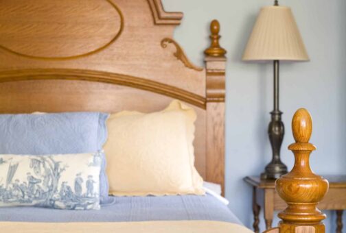 A wooden bedframe hosts a queen bed with blue and white linens with a side table and lamp in the background.